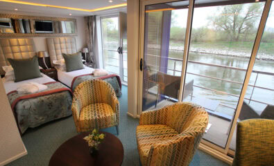 A modern AmaWaterways cabin features two single beds, a sitting area with armchairs, a small table, and a private balcony overlooking the river.