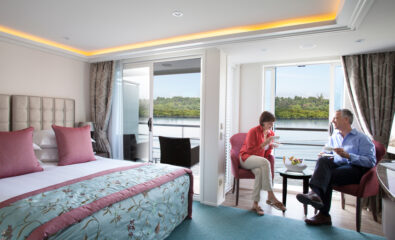 A man and woman sit by a table, drinking and talking in a bright AmaWaterways cruise ship cabin with a large bed and a stunning view of the water through open balcony doors.